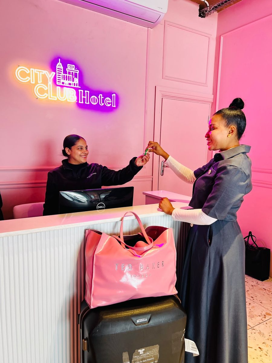 A City Club Hotel concierge handing a room key to a smiling guest at the pink reception desk, beneath the neon City Club Hotel sign